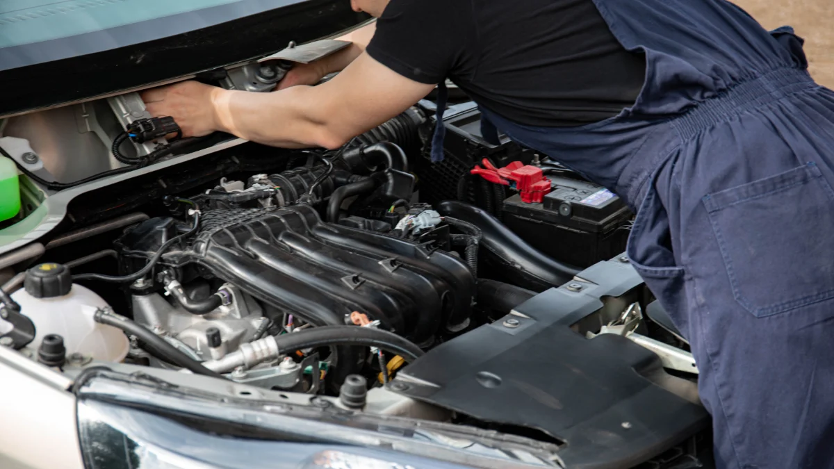 Mechanik w niebieskim kombinezonie pracuje przy silniku samochodu z otwartą maską na świeżym powietrzu w ciągu dnia, w środowisku