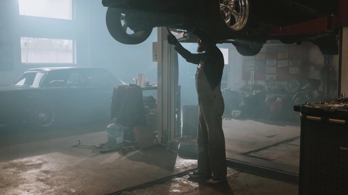 Mechanik naprawia samochód uniesiony na podnośniku w słabo oświetlonym warsztacie, tworząc vintage atmosferę, co wskazuje na złożoność i