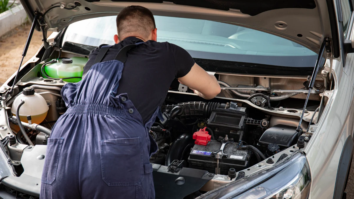 Mechanik w kombinezonie pracuje nad silnikiem samochodu na zewnątrz, prezentując umiejętności naprawy pojazdów, co jest związane z kontrolą