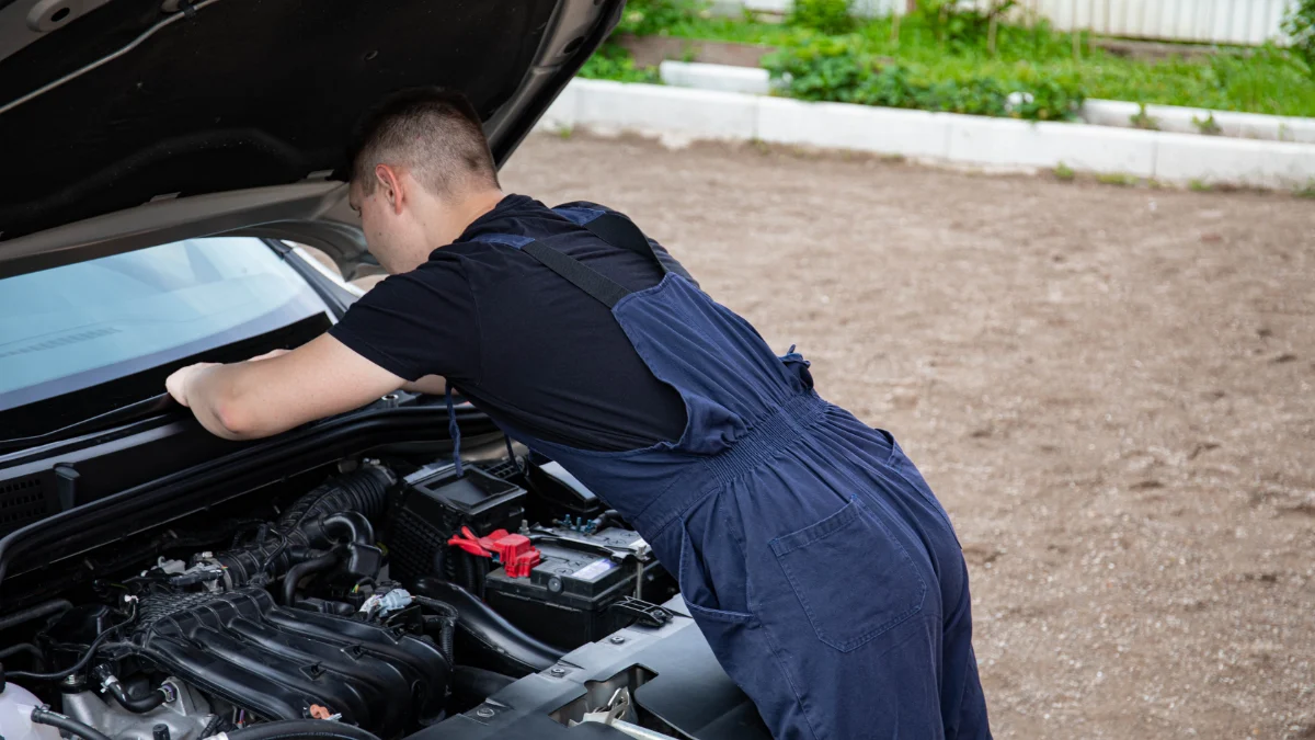 Mechanik w niebieskim kombinezonie pracuje nad naprawą silnika samochodu na świeżym powietrzu, skupiony na pracy