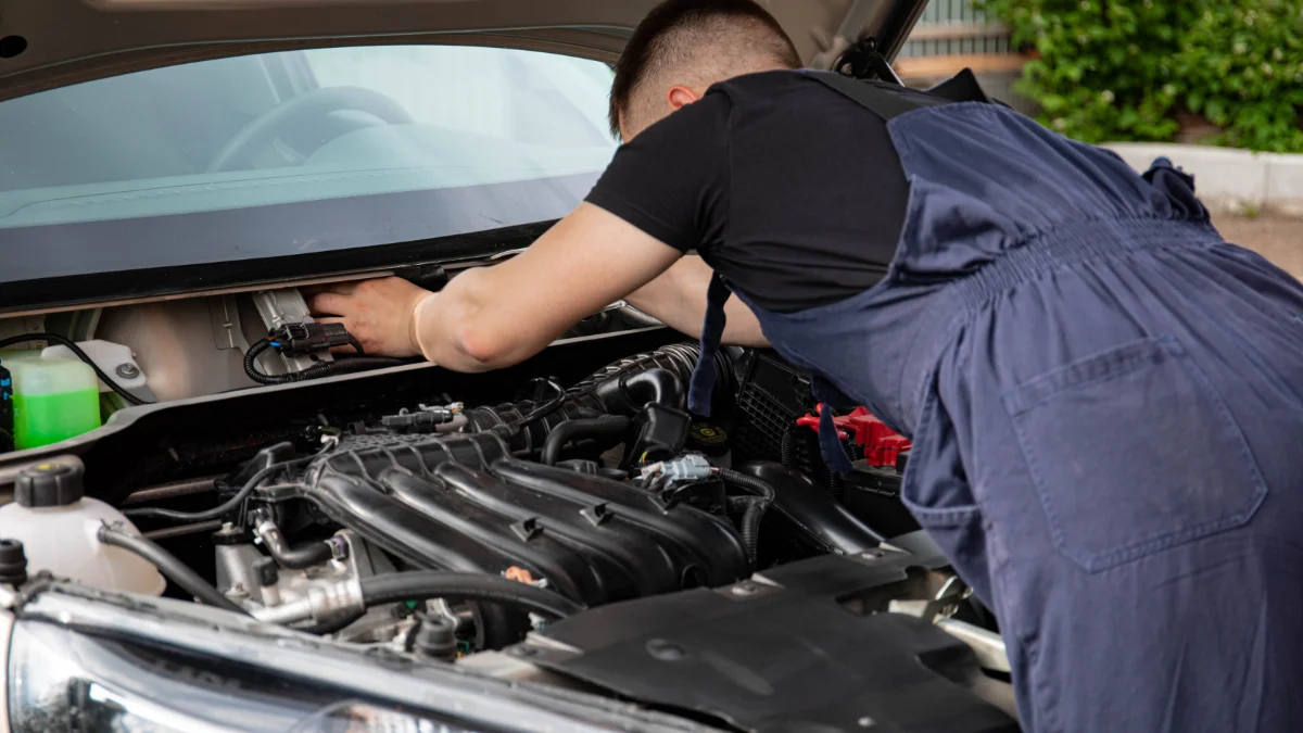 Mechanik pracuje nad serwisowaniem silnika samochodu na świeżym powietrzu, w tle widoczna jest stacja serwisowa z wyświetlonymi tablicami
